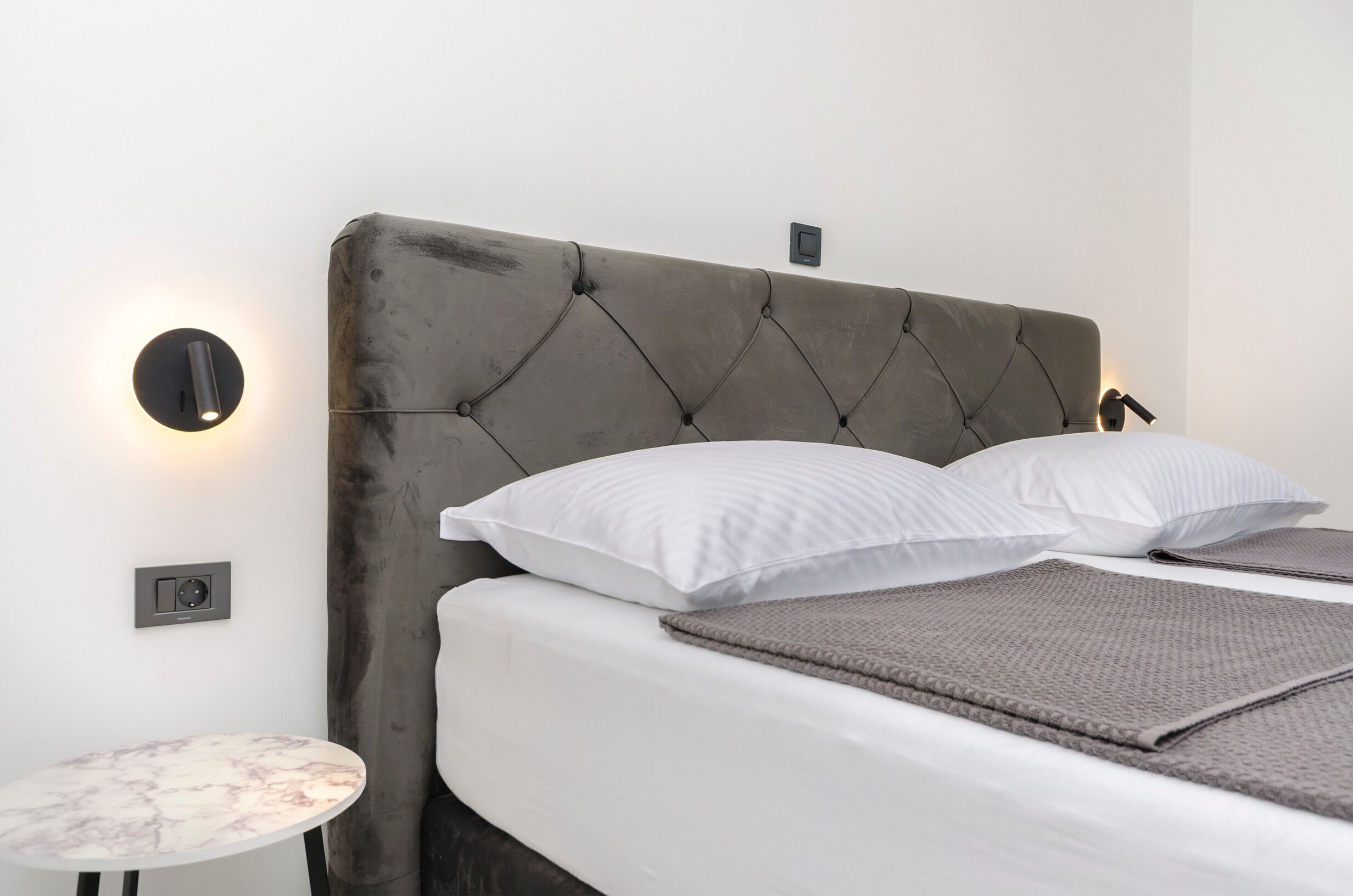 Ein ordentlich bezogenes Bett mit einem gebügelten, gepolsterten Kopfteil, zwei weißen Kissen und einer grauen Tagesdecke. Neben dem Bett steht ein kleiner runder Tisch mit Marmorplatte.