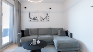 Una sala de estar moderna con un sofá seccional gris, una mesa de centro negra redonda, arte abstracto en la pared y un ventanal con cortinas.