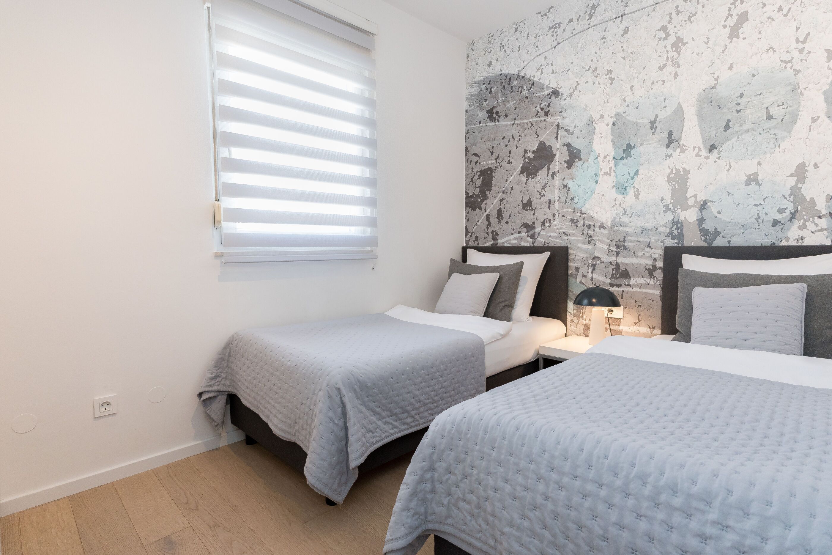 Een slaapkamer met twee bedden, een raam met zonwering, een muur met een reliëf muurschildering en een bedlampje.