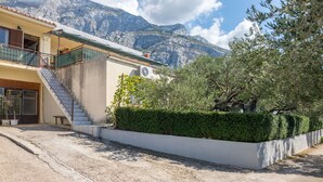 Un bâtiment à deux étages, doté d’un escalier menant à un balcon, entouré d’une haie bien entretenue et avec une chaîne de montagnes en arrière-plan.
