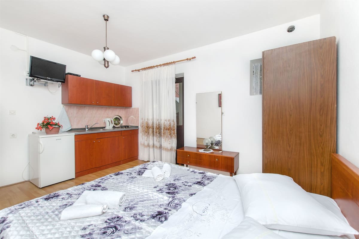 A hotel room with a bed, a wooden wardrobe, a small kitchenette, a TV, and a mirror.