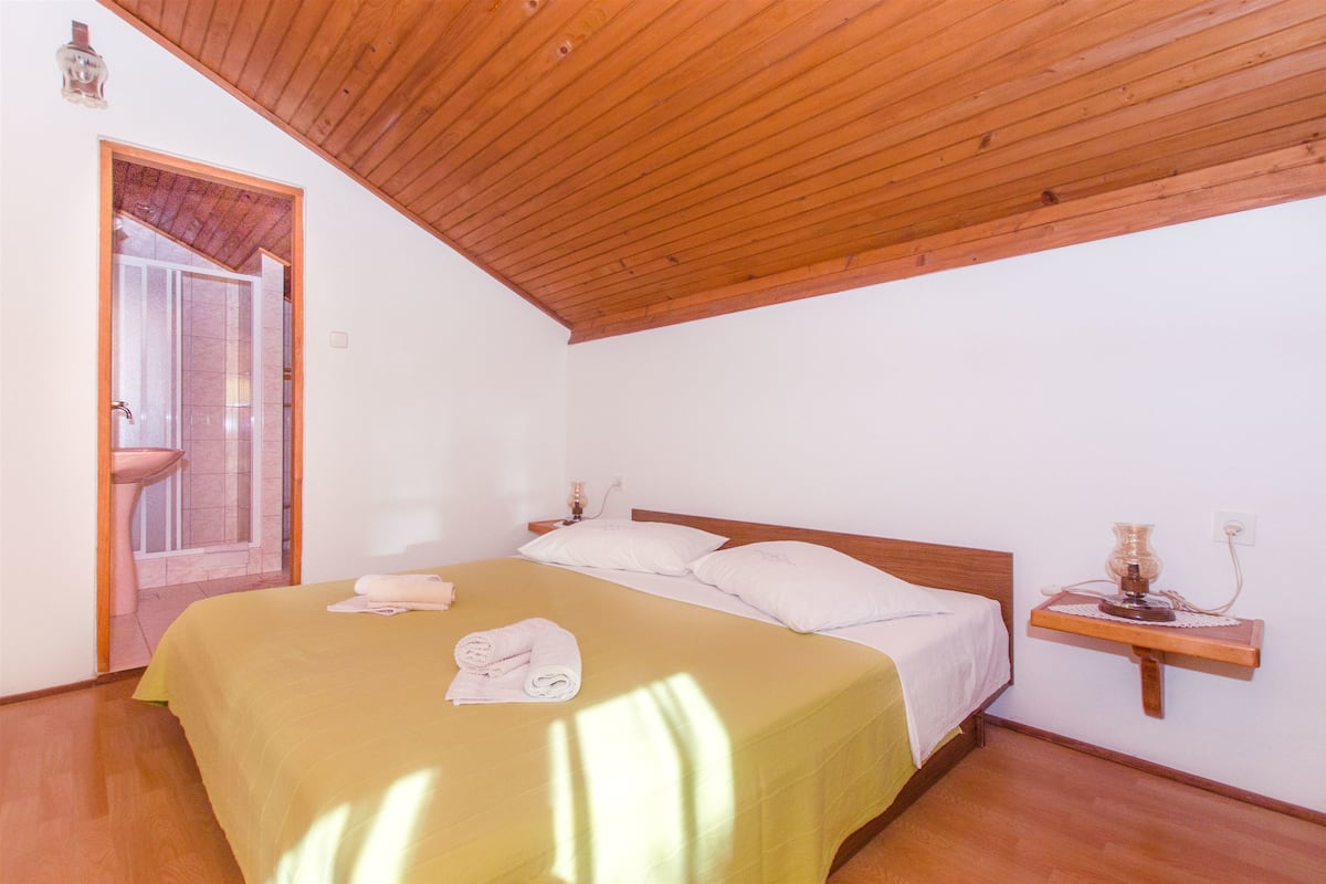 A bedroom with a wooden ceiling, a bed with a yellow bedspread, white pillows, and a bedside table with a lamp.