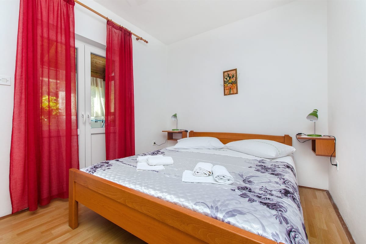 A neatly made bed with a floral bedspread and two pillows, a wooden bedside table with a green lamp, and a red curtain on the window.