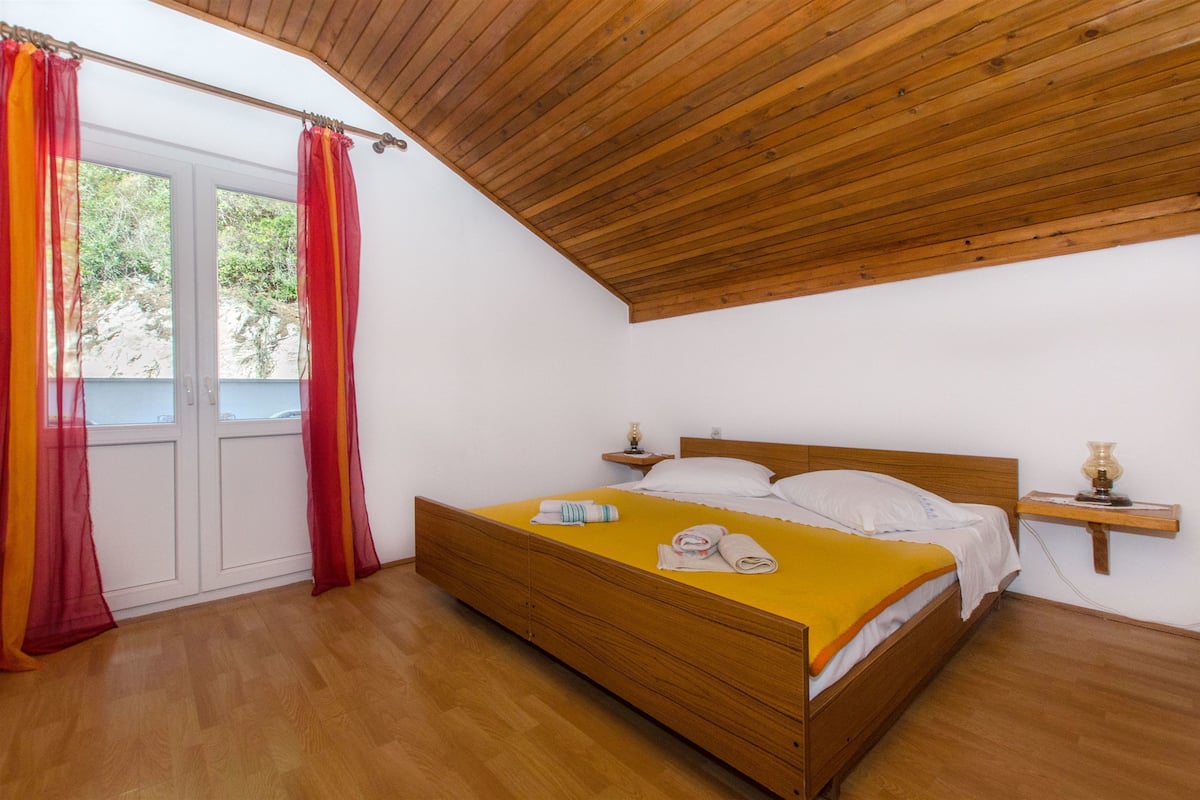 A bedroom with a wooden bed, a small bedside table, and a window with curtains.