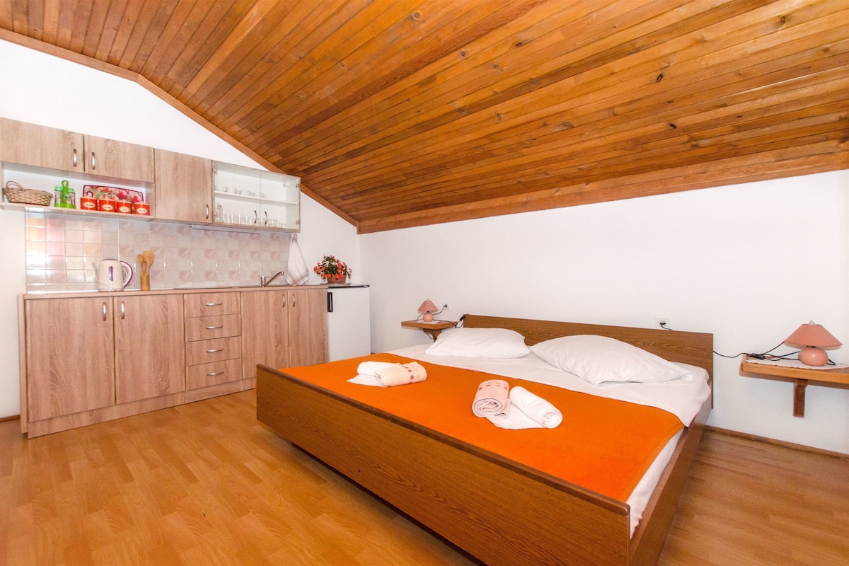 A bedroom with a wooden ceiling, a bed with an orange bedspread, a wooden wardrobe, and a small kitchen area with cabinets and a countertop.