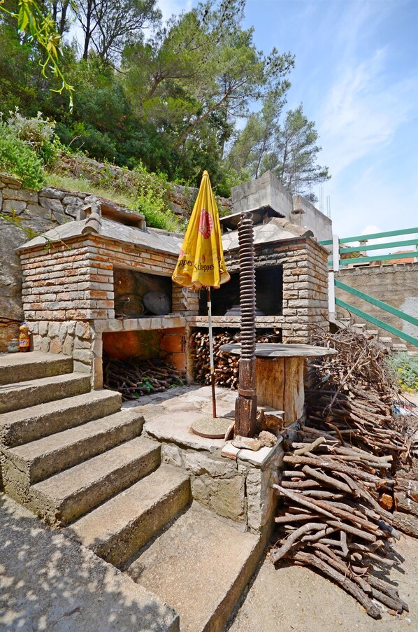 A rustic stone building with a brick oven, steps leading up to it, and a pile of firewood.