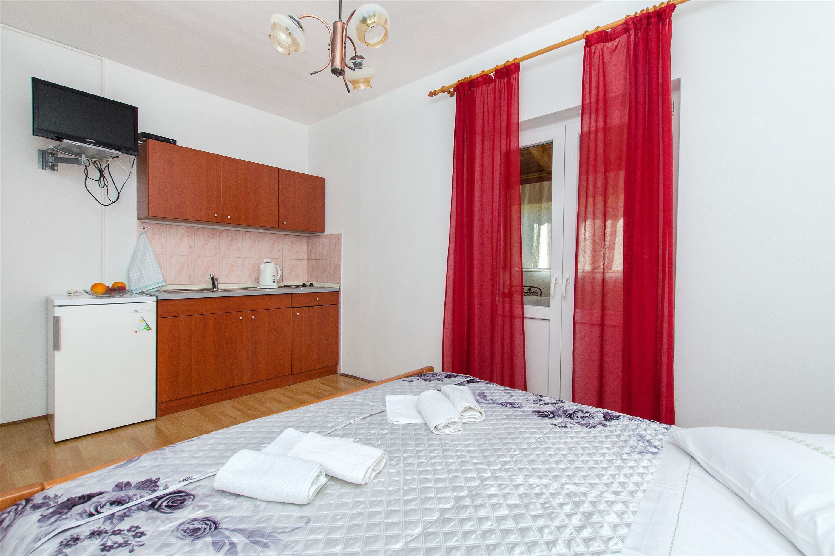 A bedroom with a bed, a small kitchenette, and a TV mounted on the wall.