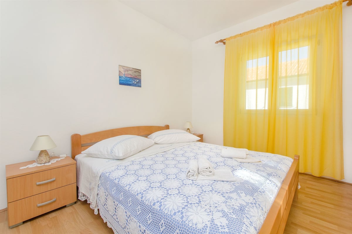 A neatly made bed with a blue and white patterned bedspread, a wooden nightstand with a lamp, and a painting on the wall.