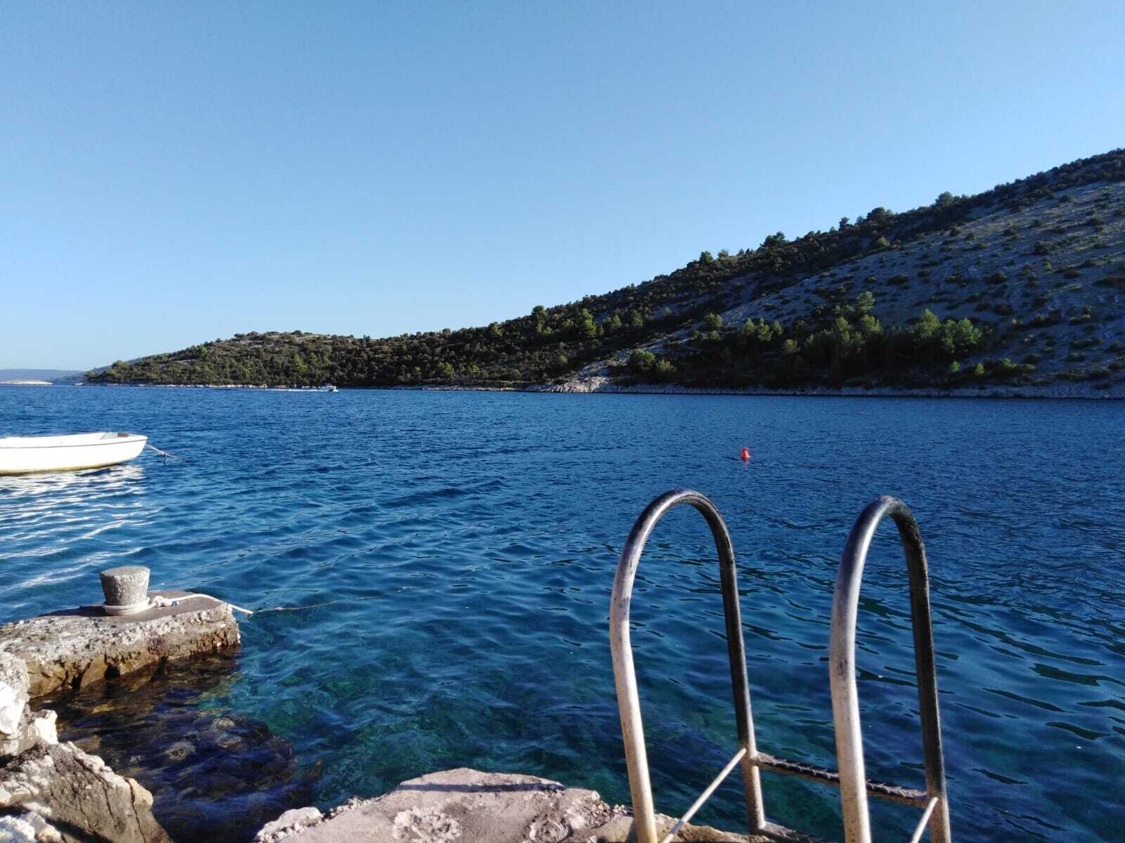 A clear blue sea with a boat docked near a rocky shore, a metal ladder, and a small island with trees in the background.