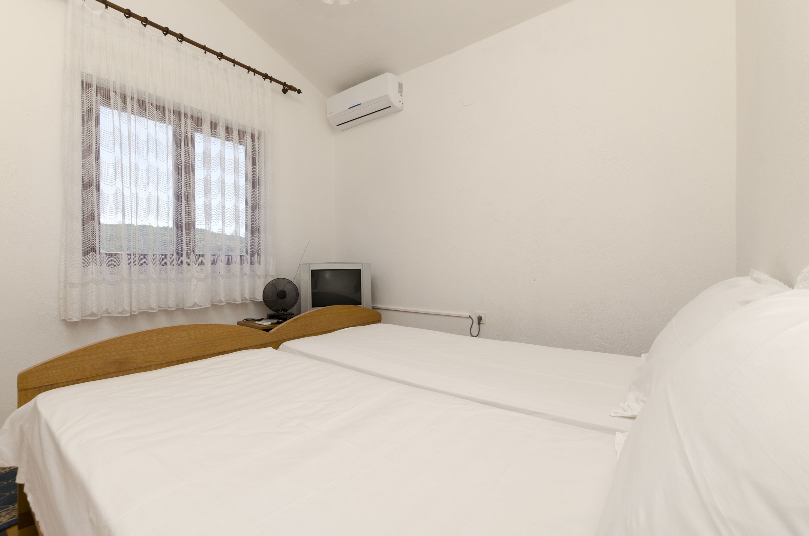 Un dormitorio con dos camas, televisión, aire acondicionado y ventana con cortinas traslúcidas.