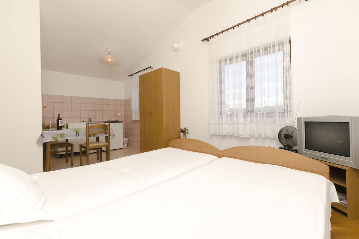 A bedroom with a bed, a TV, a wooden wardrobe, a small table with chairs, and a window with curtains.
