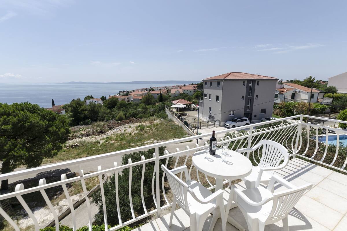 A balcony with white chairs and a table, overlooking a coastal town with buildings and a clear sky.