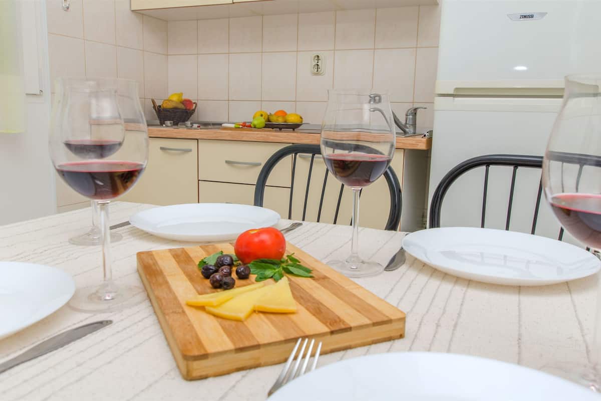 Una mesa de comedor dispuesta con platos, cubiertos, copas de vino y una tabla de madera con queso, aceitunas y tomate.