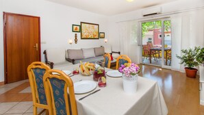 A dining area with a table set for four, chairs, a potted plant, and a sofa in the background. - Apartments Anita (Vrboska)