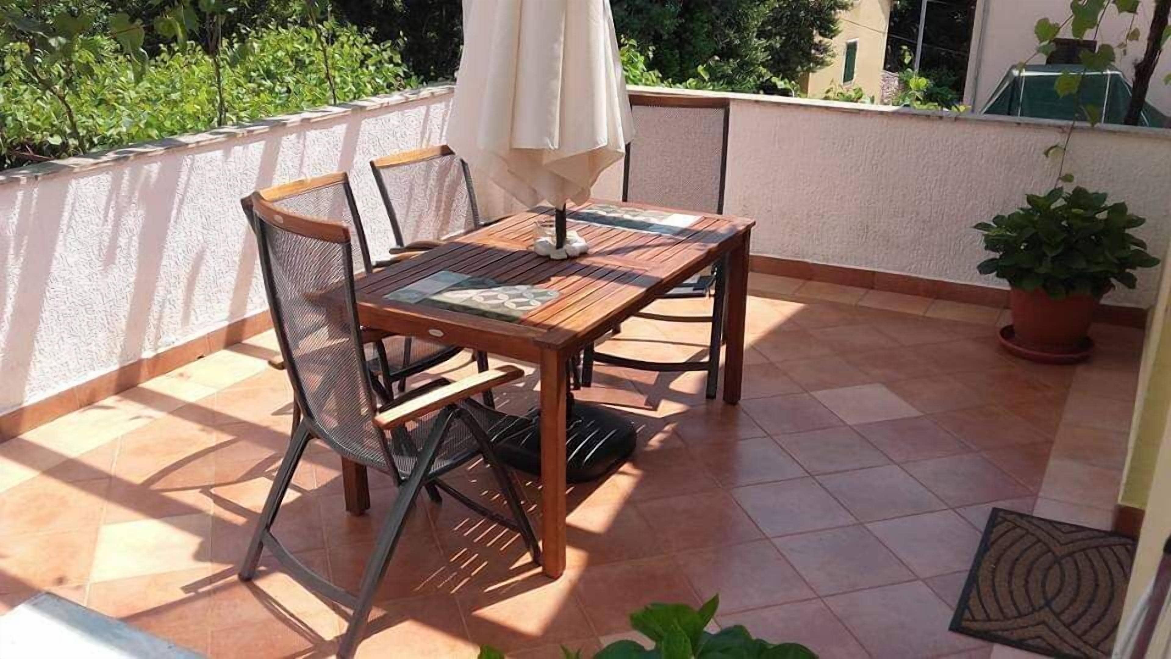 A wooden table and chairs on a tiled patio with a white umbrella, surrounded by greenery.