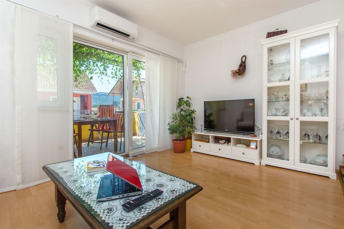 Una sala de estar con una mesa de vidrio, televisor, aparador con puertas de vidrio y una planta en maceta.
