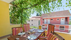 An outdoor dining area with a wooden table set for two, striped chairs, and a view of a red building in the background. - Apartments Anita (Vrboska)