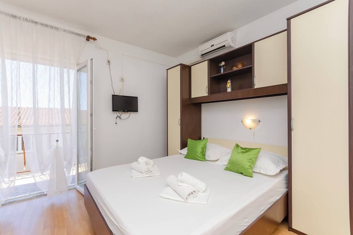 A hotel room with a bed, a TV, an air conditioning unit, and a view of the outside.