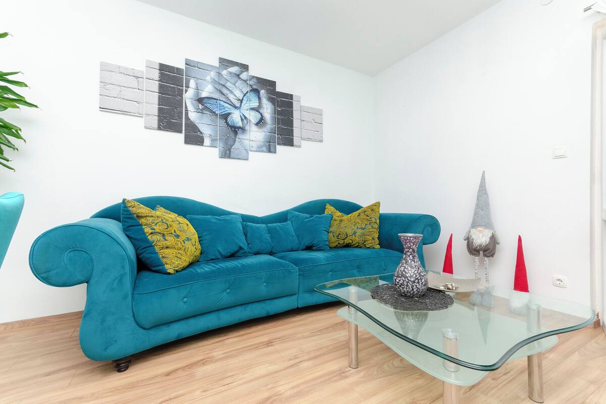 A teal sofa with patterned cushions, a glass coffee table with decorative items, and a wall-mounted gnome decoration.