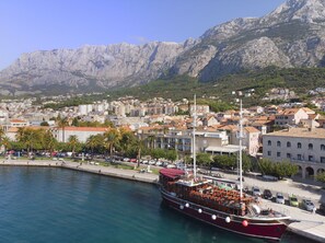 Une ville côtière où un grand navire rouge est amarré dans le port, entourée de bâtiments résidentiels et dominée par un paysage montagneux.