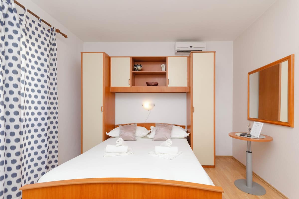 A neatly made bed with pillows, a bedside table with a lamp, a mirror, and a wardrobe with a shelf.