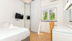 Habitación de hotel moderna con una cama, una mesa de comedor de vidrio, una silla de madera, un televisor y una ventana con vistas a zonas verdes.
