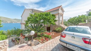 A coastal building with a car parked in front, a garden with various plants, and a clear view of the sea. - Apartments Marko (Okrug Donji)