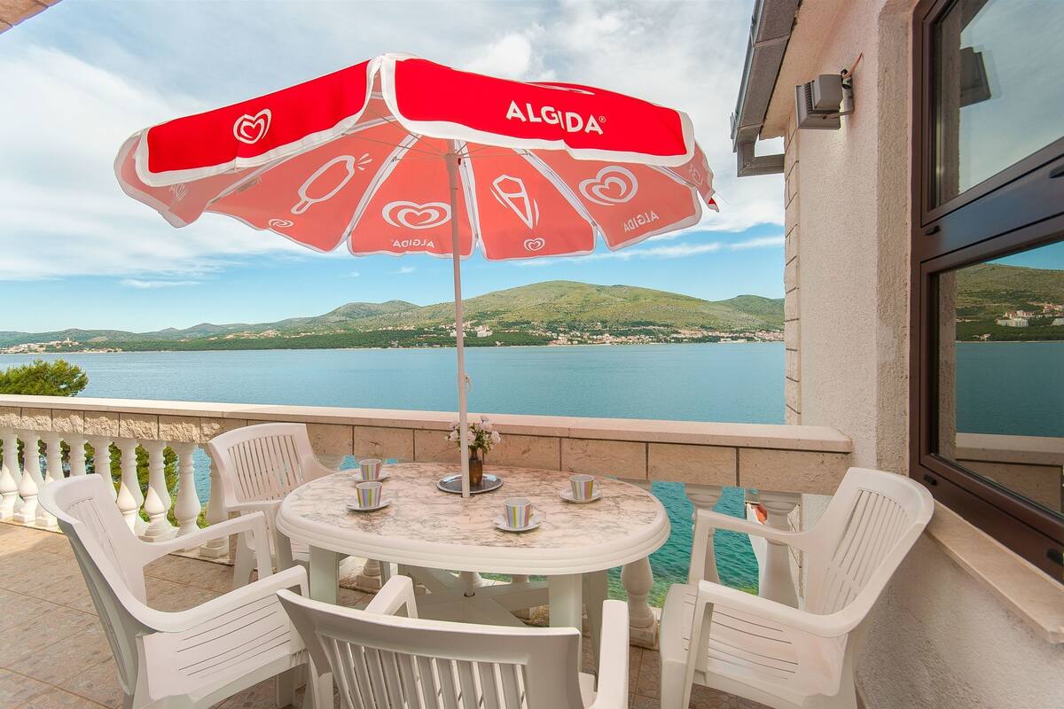 A terrace with a round table, white plastic chairs, and a red umbrella with the brand 'ALGIDA'. A view of a lake and hills is visible through the window.