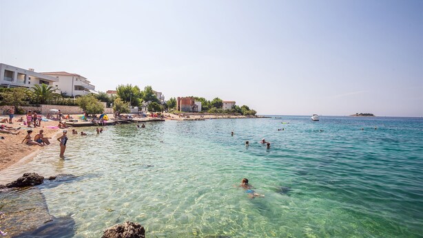 Ein Strand mit klarem Wasser, Menschen beim Schwimmen und Gebäuden im Hintergrund.