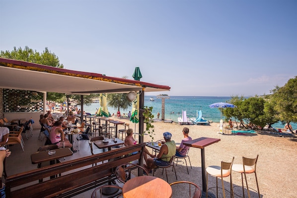 Un espace extérieur aménagé avec des tables et des chaises, donnant sur une plage parsemée de parasols et fréquentée par des gens.