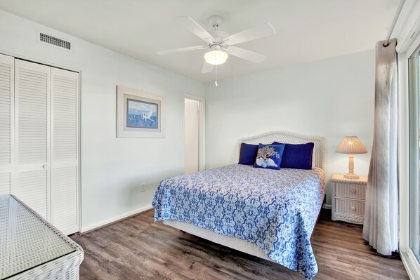 A bedroom with a bed, a ceiling fan, a nightstand with a lamp, and a wicker basket. - Lighthouse Point Condo Unit 2C (Tybee Island)