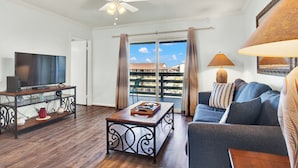 A living room with a TV, a coffee table, a sofa, a ceiling fan, and a view of a balcony with a railing. - Lighthouse Point Condo Unit 2C (Tybee Island)