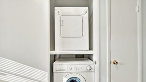 A compact laundry area with a front-loading washer and dryer, a small shelf above the dryer, and a door to the right. - Lighthouse Point Condo Unit 2C (Tybee Island)