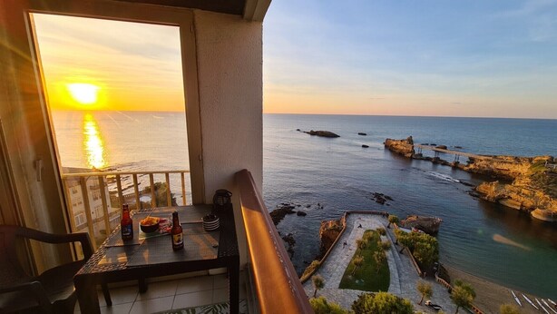 A balcony with a table set for dining, overlooking a coastal area with a rocky shoreline and a bridge. - MAGNIFICENT OCEAN VIEW BIARRITZ (Biarritz)