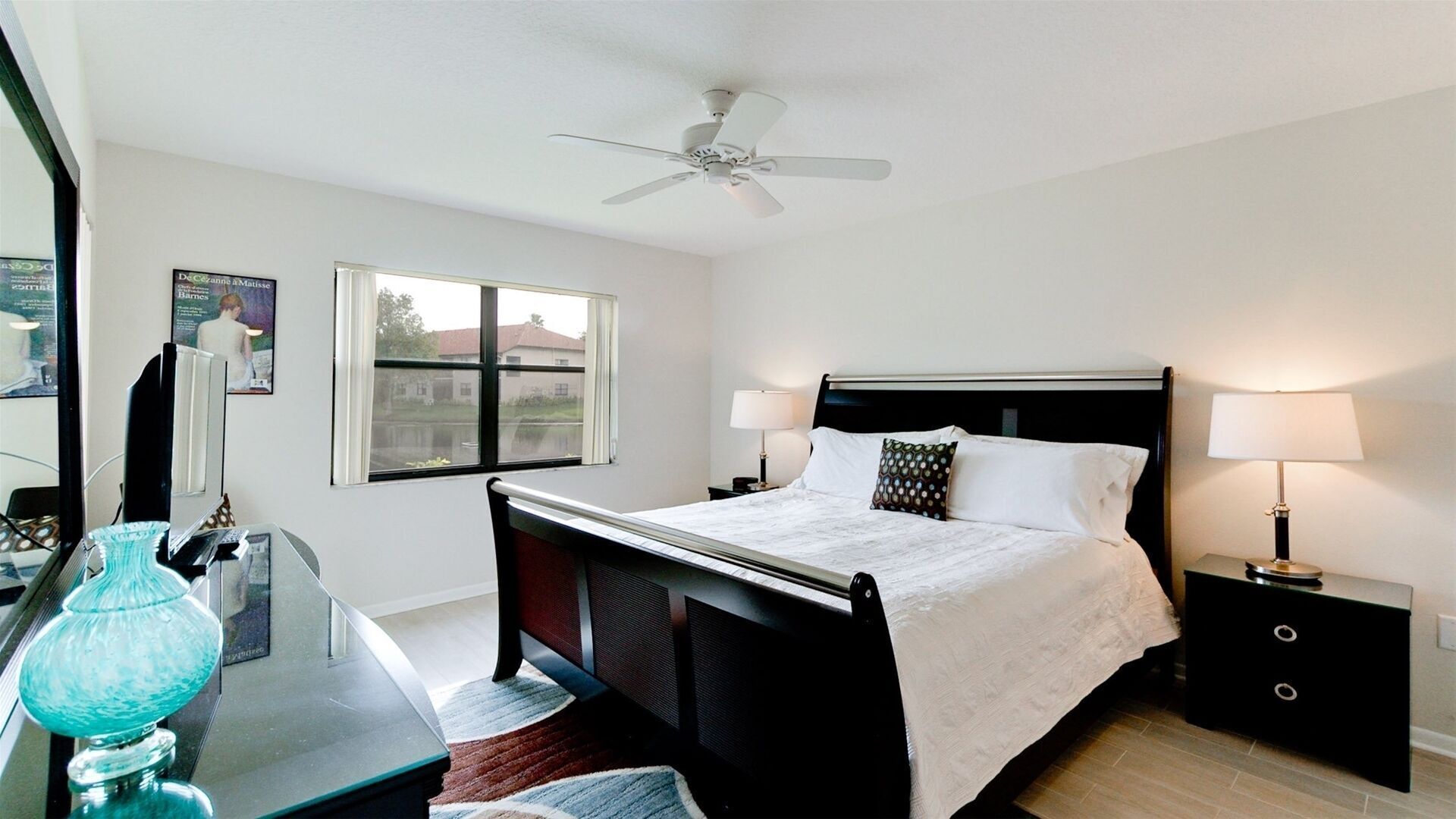 Une chambre d’hôtel avec un lit, des lampes de chevet, un bureau avec un ordinateur, un ventilateur de plafond et une fenêtre donnant sur des maisons.