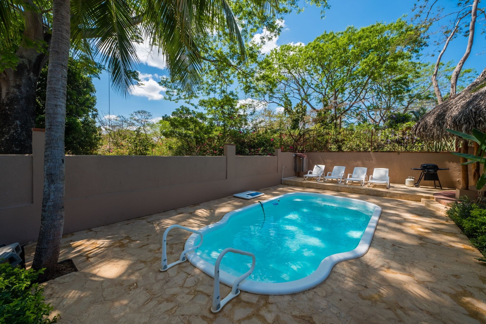 A swimming pool with a white pool ladder, surrounded by a stone patio, white lounge chairs, and a thatched roof structure.
