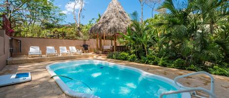 A swimming pool with a thatched-roof structure, surrounded by lush greenery and white lounge chairs.