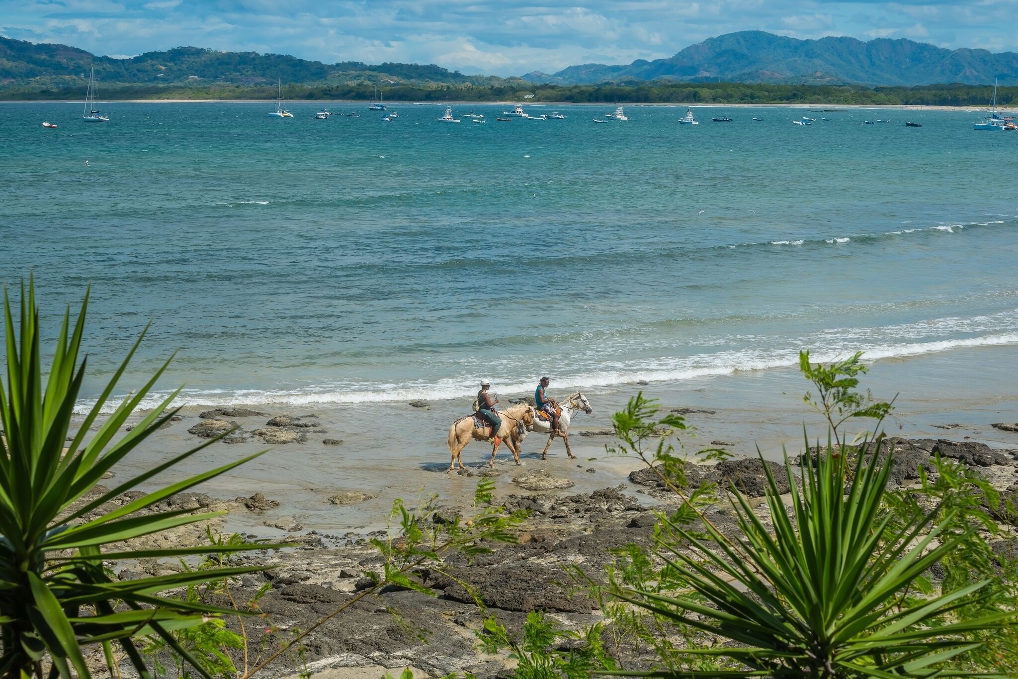 A beach with horses and riders, rocky shoreline, and boats in the water.