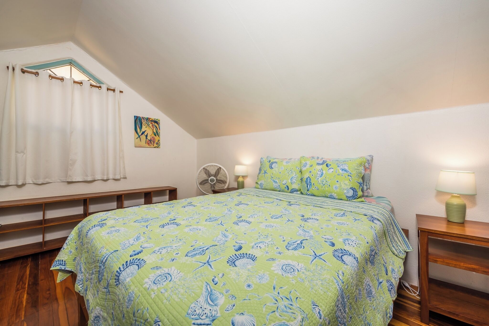 Ein Schlafzimmer mit einem Bett, einem Ventilator, einer Lampe und einem Fenster mit Vorhängen.