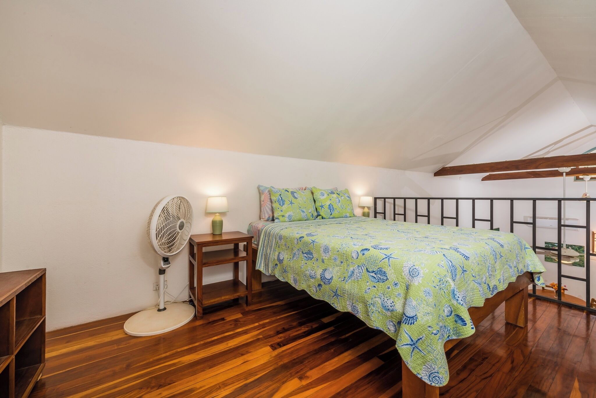 A bedroom with a wooden floor, a bed with a green patterned bedspread, a standing fan, and a nightstand with a lamp.