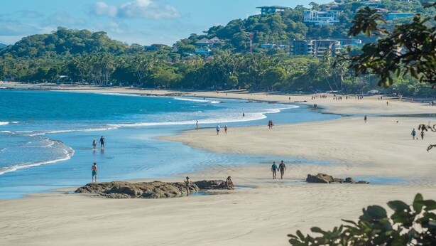 Une plage où l’on voit des gens se promener et nager, un rivage sablonneux et des bâtiments en arrière-plan.