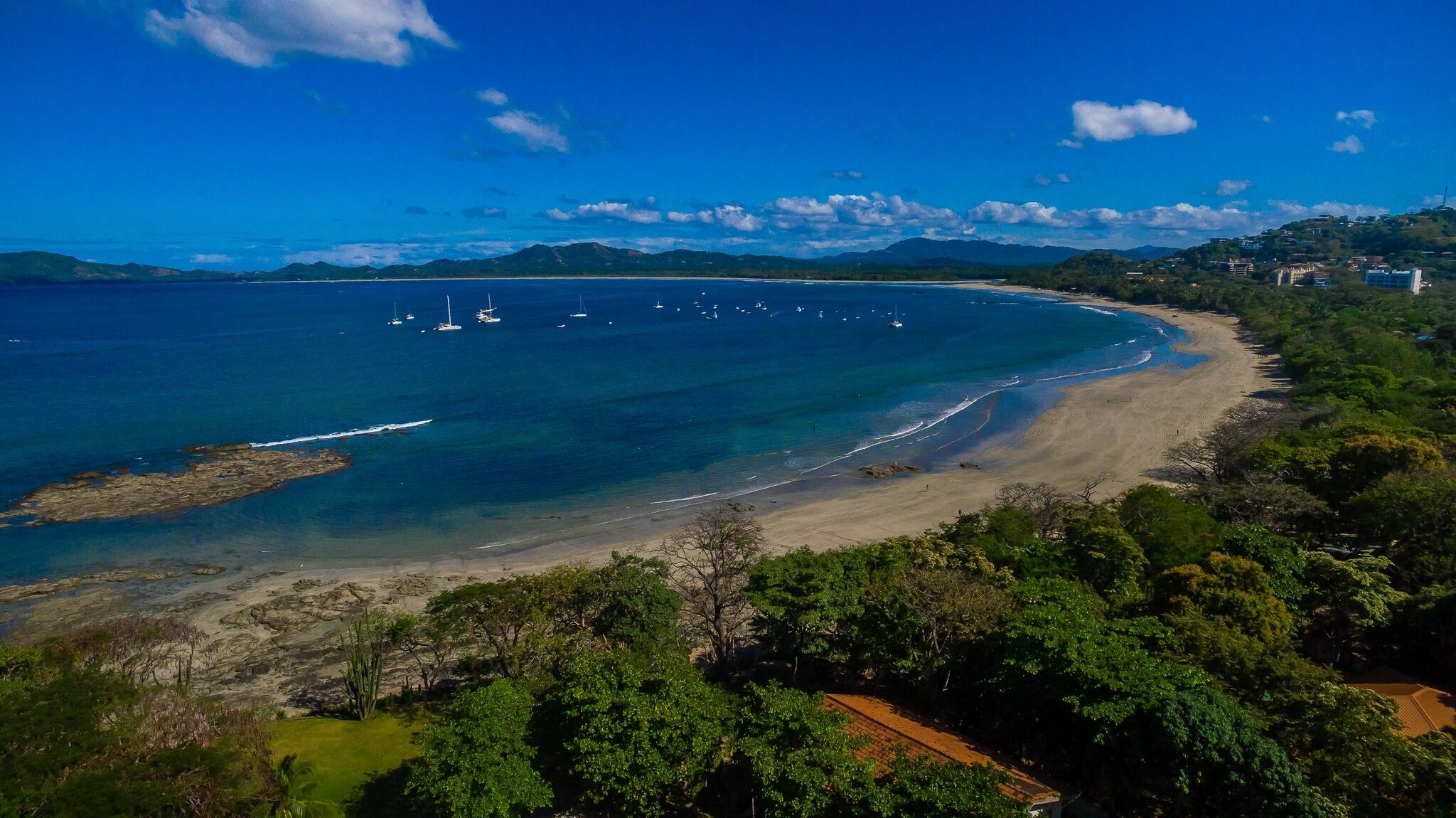 A coastal landscape with a beach, clear blue waters, and distant mountains.