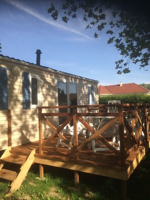 A wooden deck with a simple railing structure, a small house with a chimney, and a clear blue sky. - Camping des Nières (Ebreuil)