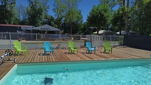 Una piscina con agua azul cristalina, rodeada por una terraza de madera. Hay varias sillas Adirondack verdes y azules, y una carpa blanca con el rótulo "LA CUINGUETTE".