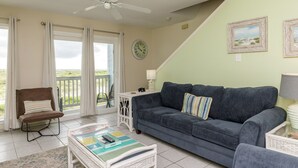 A living room with a blue sofa, a brown chair, a white table, and a ceiling fan. - Relax by the Waves | Beachside Condo with Pool & Ocean Views (Corpus Christi)