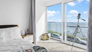 A bedroom with a bed, a nightstand, a window with a view of the sea and boats, and a telescope on the windowsill. - Lighthouse View - Woodburner & Sea View (Brixham)