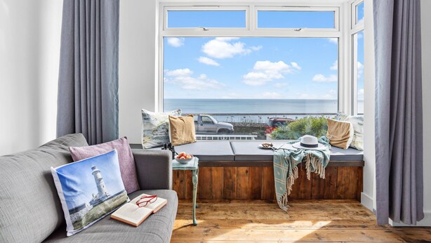 A cozy room with a large window overlooking the sea, a wooden bench with cushions, and a grey sofa with a pillow featuring a lighthouse. - Lighthouse View - Woodburner & Sea View (Brixham)