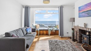A cozy living room with a grey sofa, a fireplace, and a view of the sea through large windows. - Lighthouse View - Woodburner & Sea View (Brixham)
