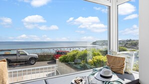 A room with a large window overlooking a coastal view, a table with a hat and a plate, and a red vehicle parked outside. - Lighthouse View - Woodburner & Sea View (Brixham)