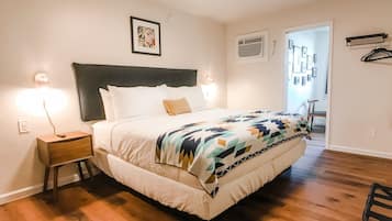 A bedroom with a bed, nightstands, a headboard, a wall-mounted air conditioner, and a door leading to another room.